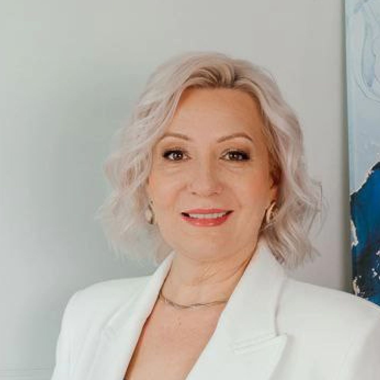 На изображении женщина с серебристо-седыми волнами и красиво уложенной прической. Она носит белый пиджак и нежный макияж, который подчеркивает ее светлую кожу и улыбающиеся глаза. У нее ярко-красная помада, которая добавляет акцент ее улыбке. Фон нейтральный, светлый с элементом современной голубой живописи, добавляющей кадру дополнительную глубину и интерес.