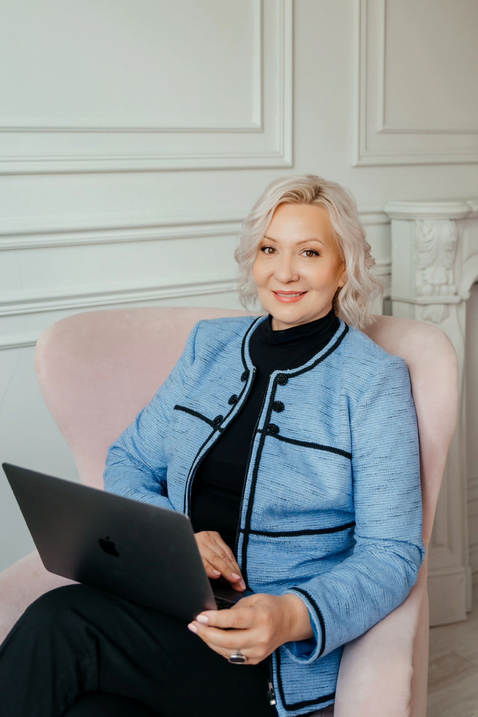 Женщина в районе 50 лет сидит на кресле и улыбается в камеру в голубом пиджаке.