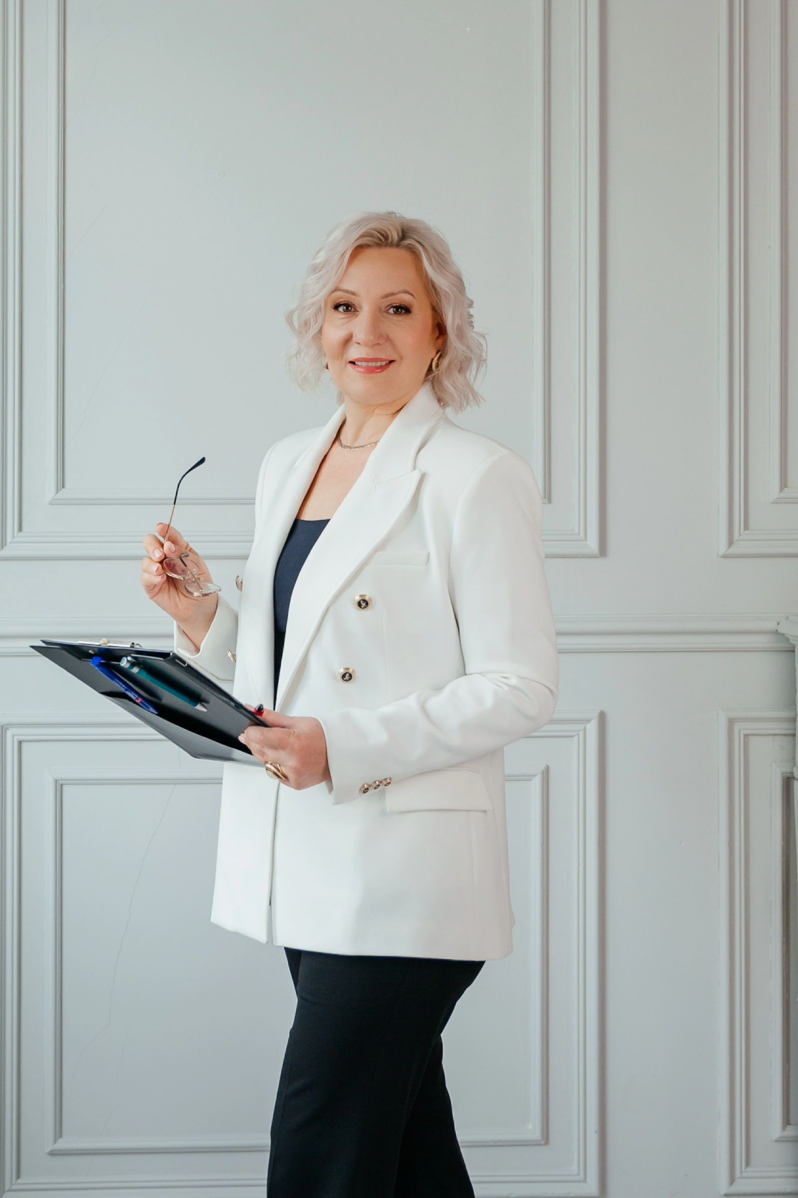 Женщина в районе 50 лет в белом пиджаке стоит и улыбается в камеру, держа в руках планшет.